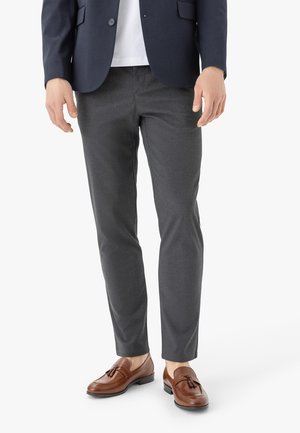 Uomo che indossa pantaloni aderenti grigio scuro, blazer blu navy, camicia bianca e mocassini marroni in pelle con nappine, in piedi su sfondo bianco.