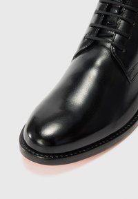 Zapato de vestir de cuero negro con acabado brillante, punta redondeada y diseño con cordones. Presenta un borde cosido a lo largo de la suela.