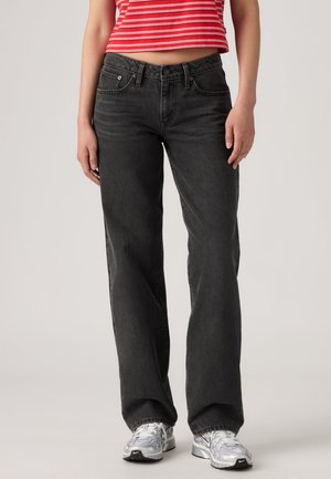 Frau trägt schwarze Jeans mit geradem Bein, weiße Sneakers und ein rot-weiß gestreiftes, bauchfreies Top und steht vor einem einfarbigen Hintergrund.