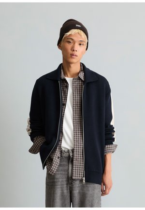 Junger Mann mit platinblonden Haaren, der eine schwarze Mütze, eine marineblaue Zip-up Jacke, ein kariertes Hemd, ein weißes T-Shirt und graue Jeans trägt und mit den Händen in den Taschen steht.