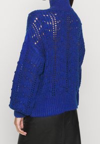 Modrý pletený svetr s vysokým límcem, volným střihem a velkými otevřenými oky. Má žebrovaný lem a široké rukávy. Doplňuje ho texturovaný vzor.
