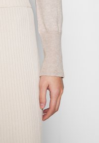 Beige ribbeadaan neuleen hihassa pehmeä rakenne, yhdistettynä vaaleampaan ribbeadaan neuleeseen. Saums yksityiskohta näkyy ranteen lähellä.