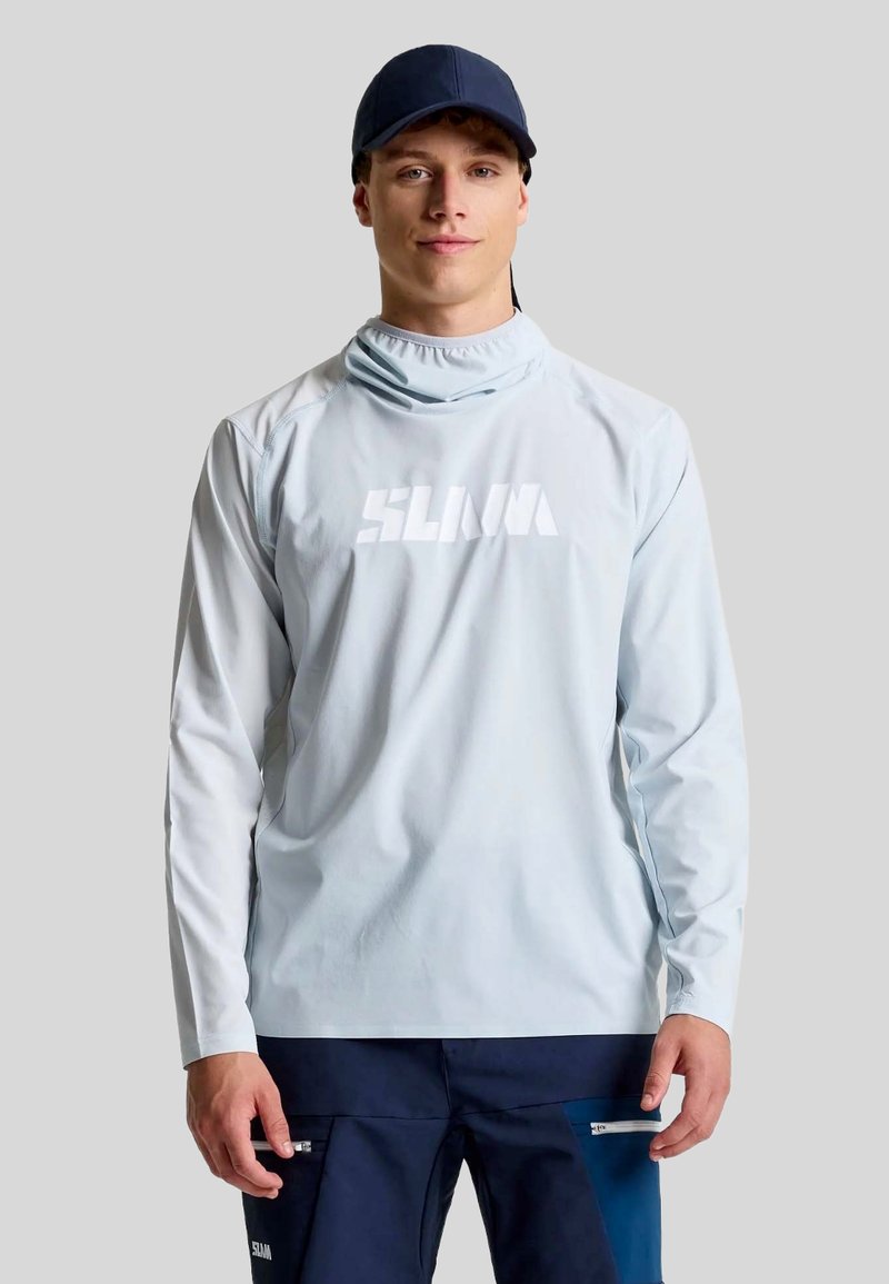Camicia azzurro chiaro a maniche lunghe con colletto alto e logo bianco, abbinata a pantaloni scuri con tasche con zip, indossata da un modello maschile.