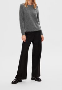 Harmaa neulepaita, jossa on pyöreä pääntie, pehmeä koostumus ja hieman rispaantunut helma, yhdistetty leveälahkeisiin mustiin housuihin ja massiivisiin mustiin kenkiin.