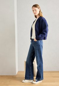 Marine bomberjakke, cremet striktop, vide blå jeans og hvide sneakers. Neutral baggrund med trægulv og en taske.