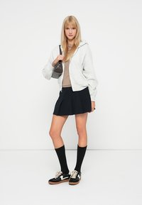 Felpa grigia con zip sopra una maglietta marrone, gonna nera plissettata, calze al ginocchio nere e sneakers nere con dettagli bianchi. Piccola borsa.