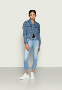 Chaqueta de mezclilla con un corte corto, dos bolsillos en el pecho, color azul claro desgastado, combinada con jeans skinny azul claro y zapatillas blancas.