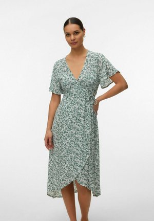 Femme portant une robe portefeuille à fleurs vertes et blanches à manches courtes, debout une main sur la hanche devant un fond uni.