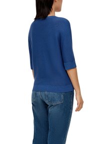 Blauer Strickpullover mit kurzen Ärmeln und einem gerippten Saum, der ein strukturiertes Muster aufweist. In Kombination mit blauen Jeans, die eine lockere Passform zeigen.