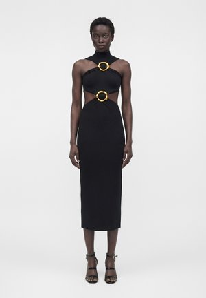 Nő fekete ujjatlan midi ruhát visel arany gyűrűs kivágásokkal a mellkasnál és a deréknál, fekete magas sarkú szandálokkal párosítva.