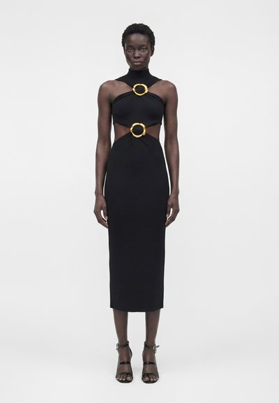 Woman wearing a black sleeveless midi dress with gold ring cutouts at chest and waist, paired with black high-heeled sandals.