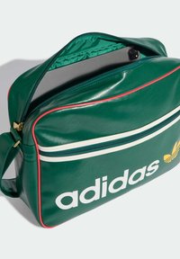 Zielona torba ze skóry syntetycznej z białymi i czerwonymi akcentami, z wyraźnym białym logo „adidas” oraz zamykanym na zamek górnym kieszonką.