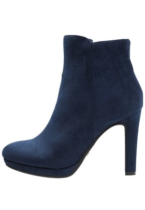 Marineblå ruskindsankelstøvle med høj blokhæl, rundet tå og sidelukning med lynlås på hvid baggrund.
