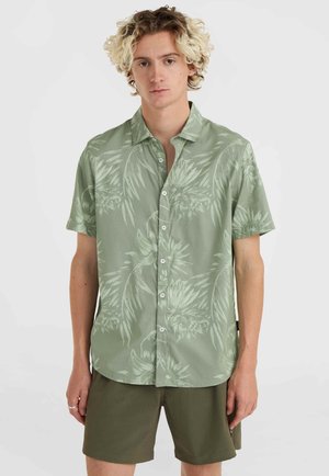 Camisa de manga corta de tela verde claro con un patrón floral. Presenta un cuello con hebilla y botones al frente. Combinada con pantalones cortos de color verde oliva.