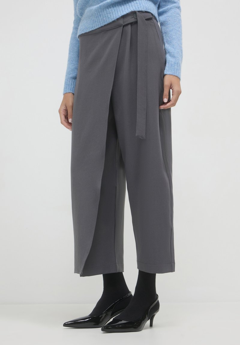 Femme portant un pantalon large gris en wrap, des collants noirs et des chaussures à talon kitten en cuir verni noir, se tenant contre un fond blanc.