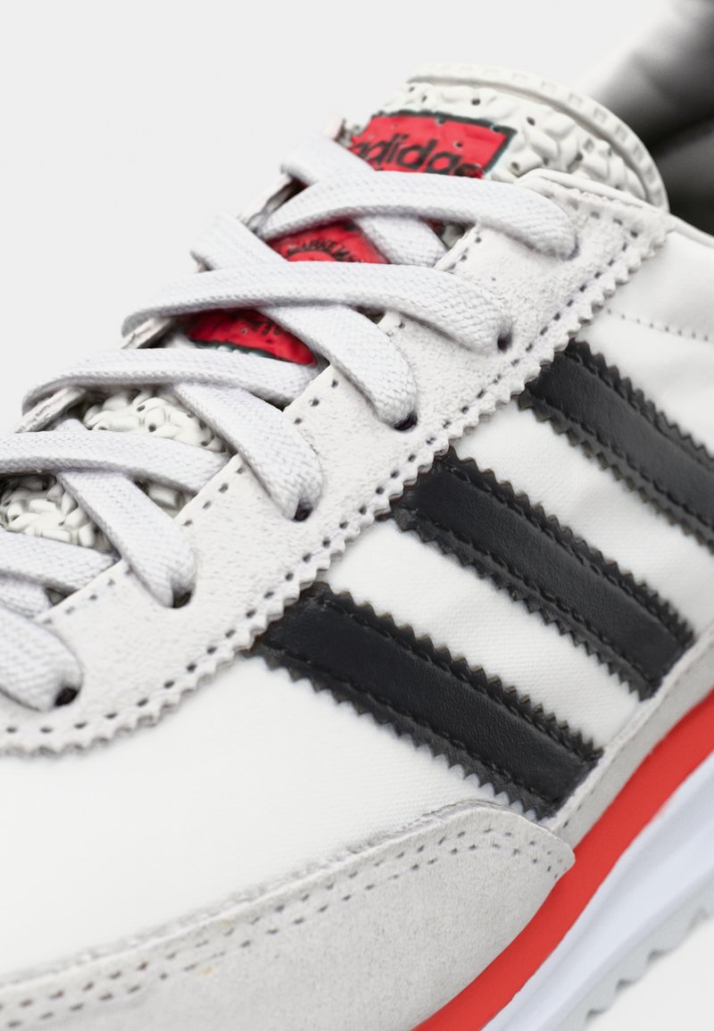 Chaussure de sport blanche en cuir et textile, avec des rayures noires, des accents gris et une étiquette de logo rouge près du col rembourré.