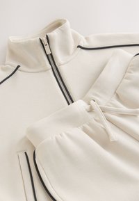 Cream-colored sweatshirt and pants set, featuring a mock neck, zip closure, and black piping accents. Soft, textured fabric.