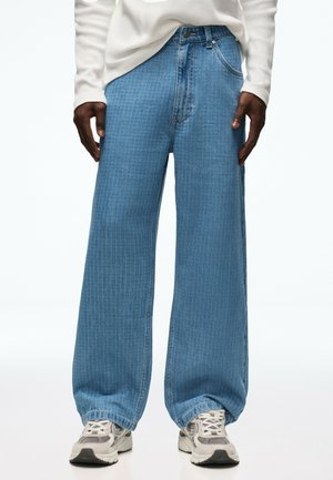 Personne portant un jean bleu à jambes larges avec un motif en grille subtil, des baskets blanches et un haut blanc à manches longues, debout devant un fond blanc.