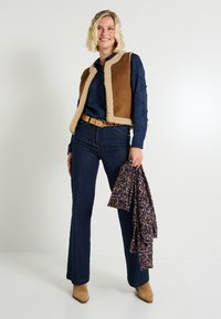 Mujer sonriendo, vestida con una camisa bordada azul marino, chaleco de pelo sintético marrón, jeans oscuros, cinturón y botas marrones, sosteniendo una tela floral con la mano derecha.
