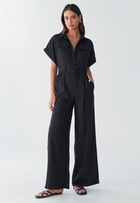Mono negro hecho de tejido suave, con botones en la parte delantera, mangas cortas, dos bolsillos en el pecho, cintura ceñida con cinturón y pantalones de pierna ancha.