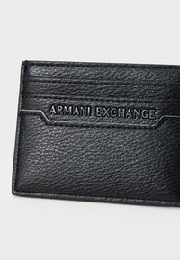 Juodas odinis kortelių laikiklis su įspaustu "ARMANI EXCHANGE" logotipu. Siūlo tekstūruotą paviršių ir kontrastingus siūlus, suteikiančius papildomų detalių.