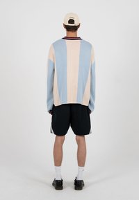 Homme de dos portant un pull beige et bleu à rayures, un short noir, des chaussettes blanches, des chaussures noires et une casquette de baseball beige.