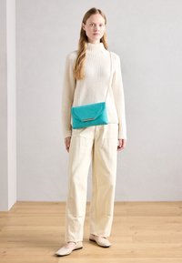 Cremfarbener gerippter Pullover, beige weit geschnittene Hose und hautfarbene Ballerinas. Eine türkisfarbene Umhängetasche mit einem goldakzentuierten Klappdeckel rundet das Outfit ab.