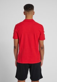 Rød bomulds polo shirt med standard krave og korte ærmer. Har en glat tekstur, sideslids og hvide logo accenter på ærmerne.