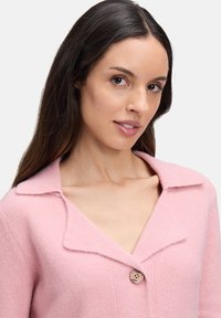 Frau mit langen dunklen Haaren, die eine geknöpfte rosa Wolljacke mit weitem Kragen vor einem schlichten hellen Hintergrund trägt.