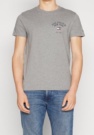 Mann trägt ein graues Tommy Hilfiger T-Shirt mit Logo und dem Schriftzug "Hilfiger New York Est. 1985" auf der Brust, kombiniert mit blauen Jeans.