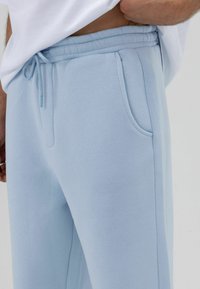 Pantalon de survêtement bleu clair avec une texture lisse, doté d'une taille élastique avec cordon, de poches avant et d'une coupe décontractée.