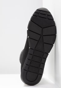 Semelle de chaussure noire avec un motif de bande de roulement texturé, inclinée contre un mur blanc et un sol blanc.