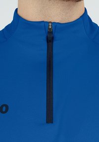 Maglione sportivo blu con zip, dotato di cerniera nera, colletto corto e una texture liscia. Include un logo stampato sul davanti.