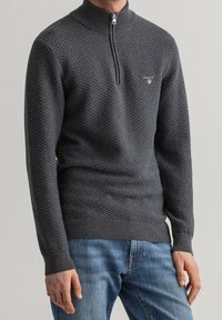 Pull en tricot gris foncé avec un col montant et une demi-fermeture éclair, présentant un motif en diamant texturé et des bords côtelés aux manches et au bas.