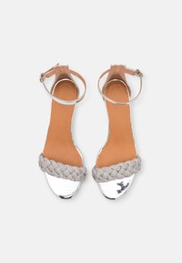 Srebrne sandały z odkrytymi palcami, z plecioną tkaninową paskiem i zapięciem na kostkę. Gładka metaliczna powierzchnia i brązowa wkładka. Płaska podeszwa.