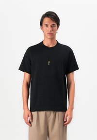 Bărbat îmbrăcat cu un tricou negru cu mânecă scurtă, având un grafic mic cu un soare și un turn pe piept și pantaloni bej, stând în fața unui fundal alb.