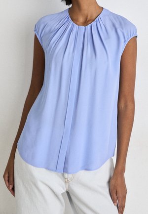 Femme portant un chemisier sans manches bleu clair avec des plis au niveau de l'encolure et un pantalon blanc sur un fond uni.