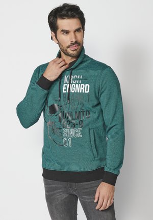Sudadera de color verde azulado con un patrón texturizado, bolsillo frontal, capucha con cordón y gráficos impresos en blanco y negro, combinada con pantalones vaqueros oscuros.