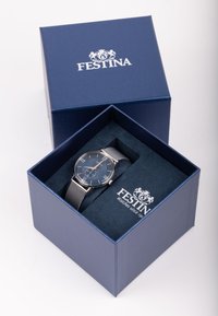 Festina rokas pulkstenis zilā kastē. Tam ir tumši zila ciparnīca, sudraba sietiņa siksna, nerūsējošā tērauda korpuss un balti stundu rādītāji. Kastei ir iekļauts logo.