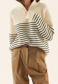 Pull crème à rayures avec des lignes horizontales noires, texture côtelée et col à demi-fermeture éclair, associé à un pantalon plissé marron avec un bouton.