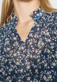 Primo piano di una blusa traslucida blu scuro con un motivo floreale, colletto arricciato e una collana doppia in argento indossata da una persona.