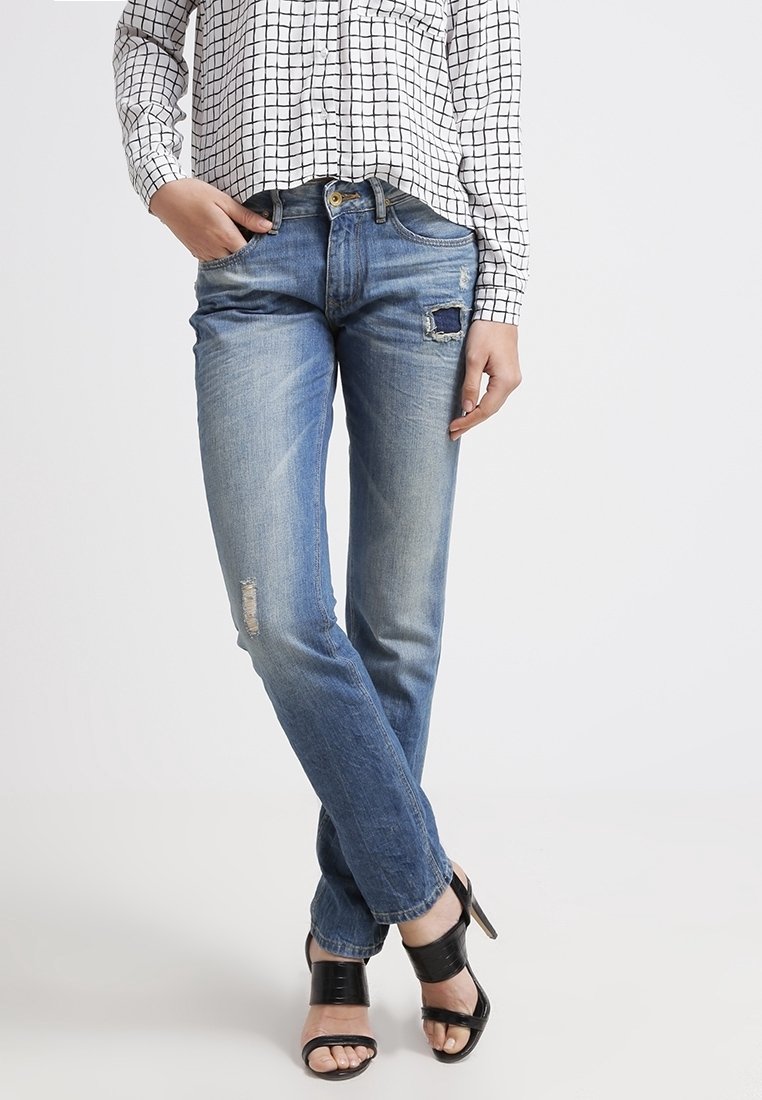 Femme portant un jean bleu clair déchiré, un chemisier blanc à motif à carreaux, et des sandales noires à talons hauts, debout avec les jambes croisées.