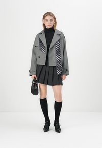 Giacca grigia corta con dettagli a pois neri, indossata sopra un dolcevita nero e una gonna grigia plissettata. Calze nere al ginocchio e scarpe.