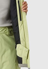 Hellgrüne Jacke mit schwarzem Innenfutter, Reißverschlusstasche und Druckknopfverschluss an den Ärmelbündchen. Glatte Textur mit modernem Design.