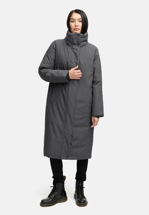 Kvinna klädd i en lång mörkgrå vadderad kappa med hög krage och svarta snörstövlar, stående framför en enfärgad vit bakgrund.