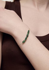 Złoty bransoletka z łańcuchem, z czterema prostokątnymi zielonymi kamieniami szlachetnymi o fakturowanej powierzchni, osadzonymi w wypolerowanych metalowych akcentach.