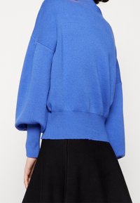 Blauer gestrickter Pullover mit hohem Kragen, überschnittenen Schultern und einem gerippten Saum. Weiche Textur mit lockerem Schnitt. Kombiniert mit einem schwarzen Rock.