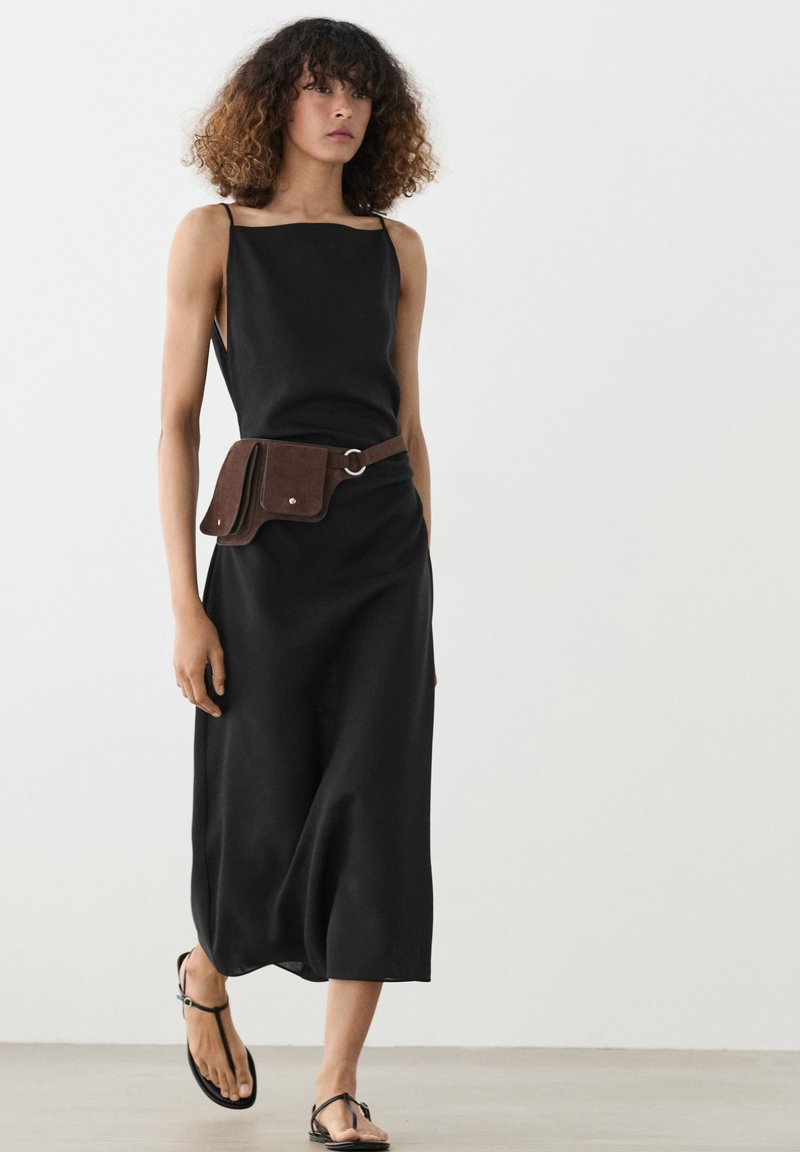 Woman in black sleeveless midi dress, wearing brown leather belt bag and black thong sandals, standing against a plain light background.