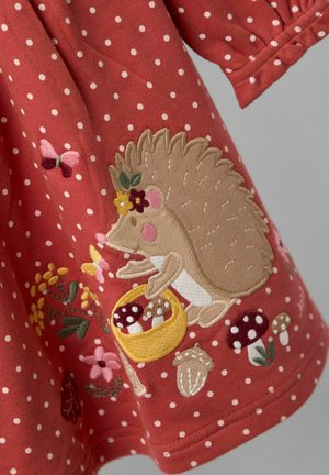 Punainen kangasmekko, jossa on valkoisia pilkkuja, ja jossa on brodeerattu siili, joka pitää sienikoria, ympärillään kukkia ja perhosia.