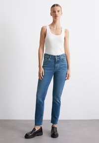 Top de tirantes blanco con canalé y jeans de tiro alto, rectos y azules con dobladillo deshilachado. Unos zapatos negros tipo slip-on completan el conjunto.